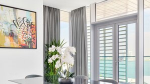 Ein moderner Essbereich mit einem Glastisch, vier Stühlen mit Holz- und Grau-Polsterung, einer Vase mit weißen Blumen und einem großen abstrakten Gemälde an der Wand.