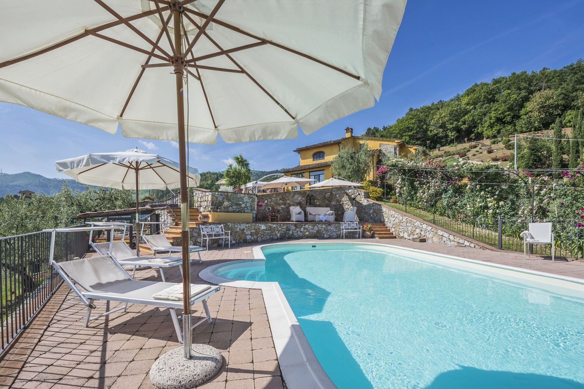 Un espace piscine agrémenté de chaises longues et de parasols, entouré de murs en pierre et offrant une vue sur une colline.