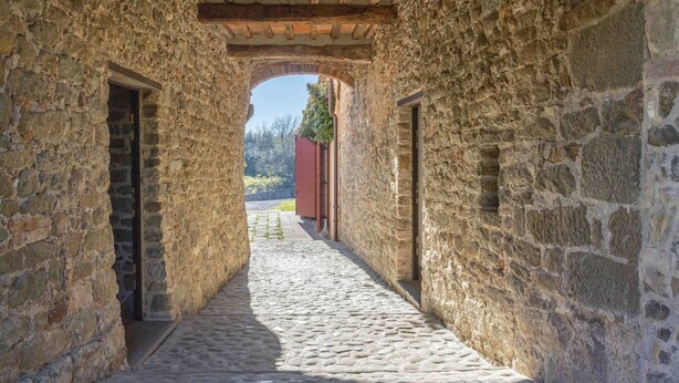 Un corredor de piedra con un sendero adoquinado, vigas de madera y un edificio rojo a lo lejos.