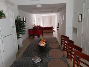Amplia sala de estar con mesa de comedor, sillas y frutero. Se observa un sofá rojo y una planta al fondo.