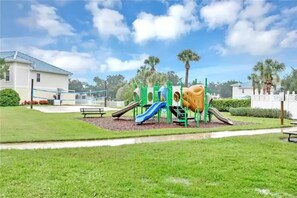 Un parque infantil con toboganes y estructuras para escalar, rodeado de un césped cuidado y un edificio al fondo.