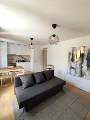 Relax in this stylish living space with a sofa, dining table, and kitchen area, featuring a unique pendant light design and an abstract art piece.