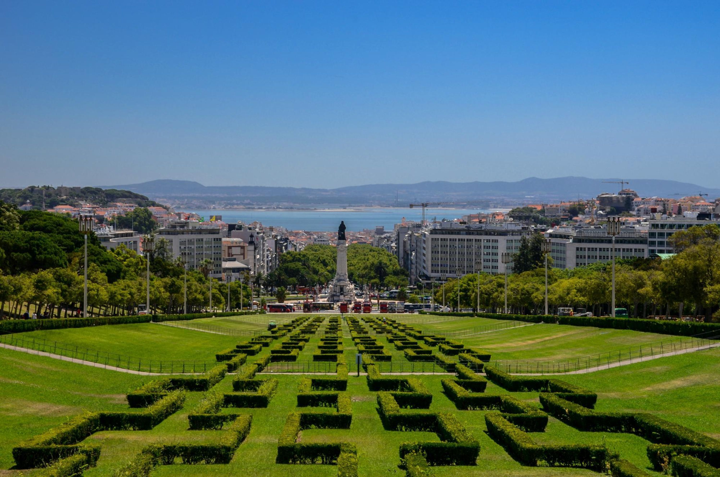 A large, open public space with a central monument, surrounded by neatly trimmed hedges and a variety of buildings in the background.