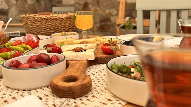 A table set with various food items, including fruits, cheeses, and a glass of orange juice, in a stone-walled outdoor dining area. - Hanole Guest House (Ayvalik)