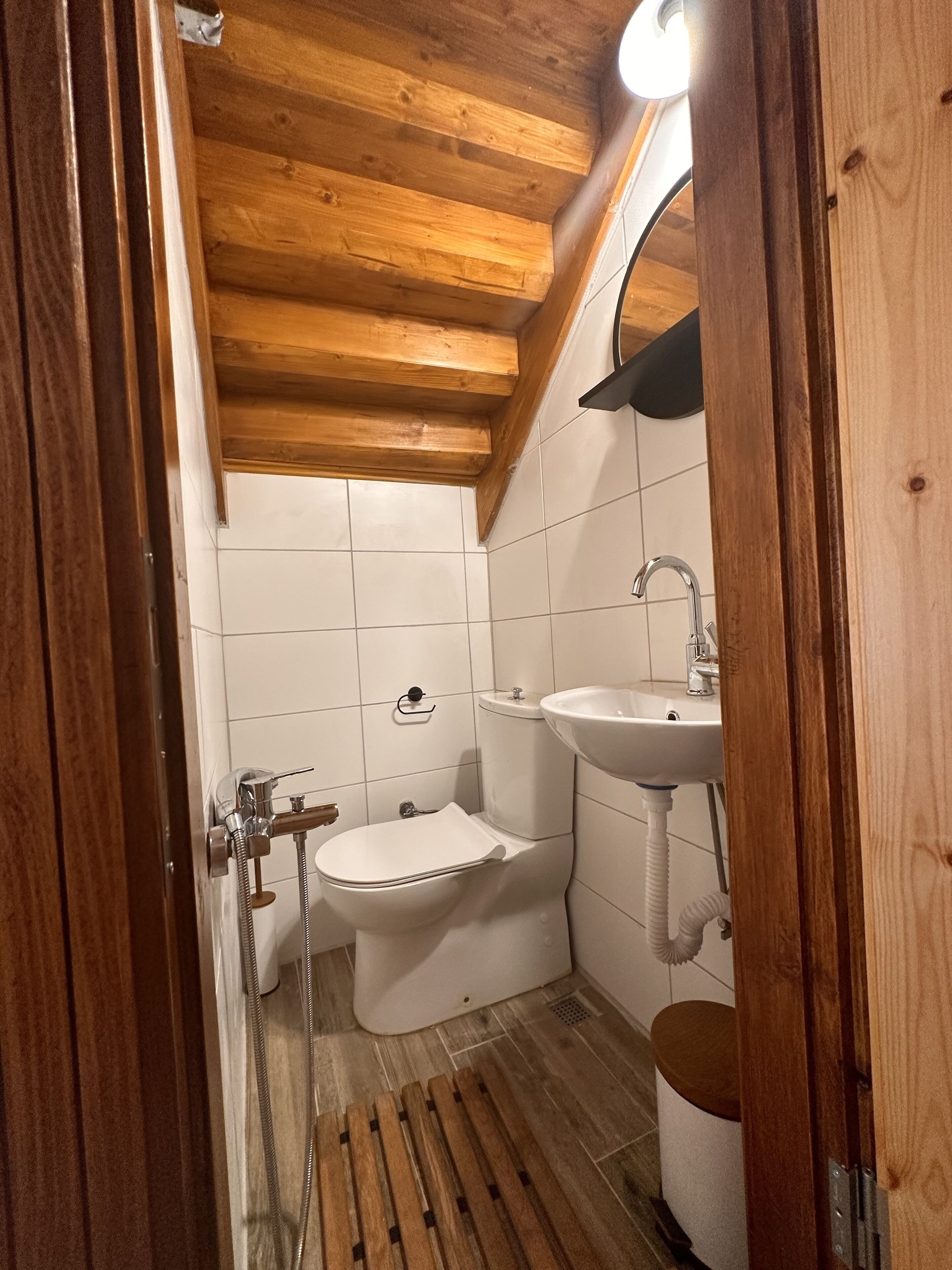 Een kleine, compacte badkamer met een houten plafond, witte tegels, een toilet, een wastafel en een douche.