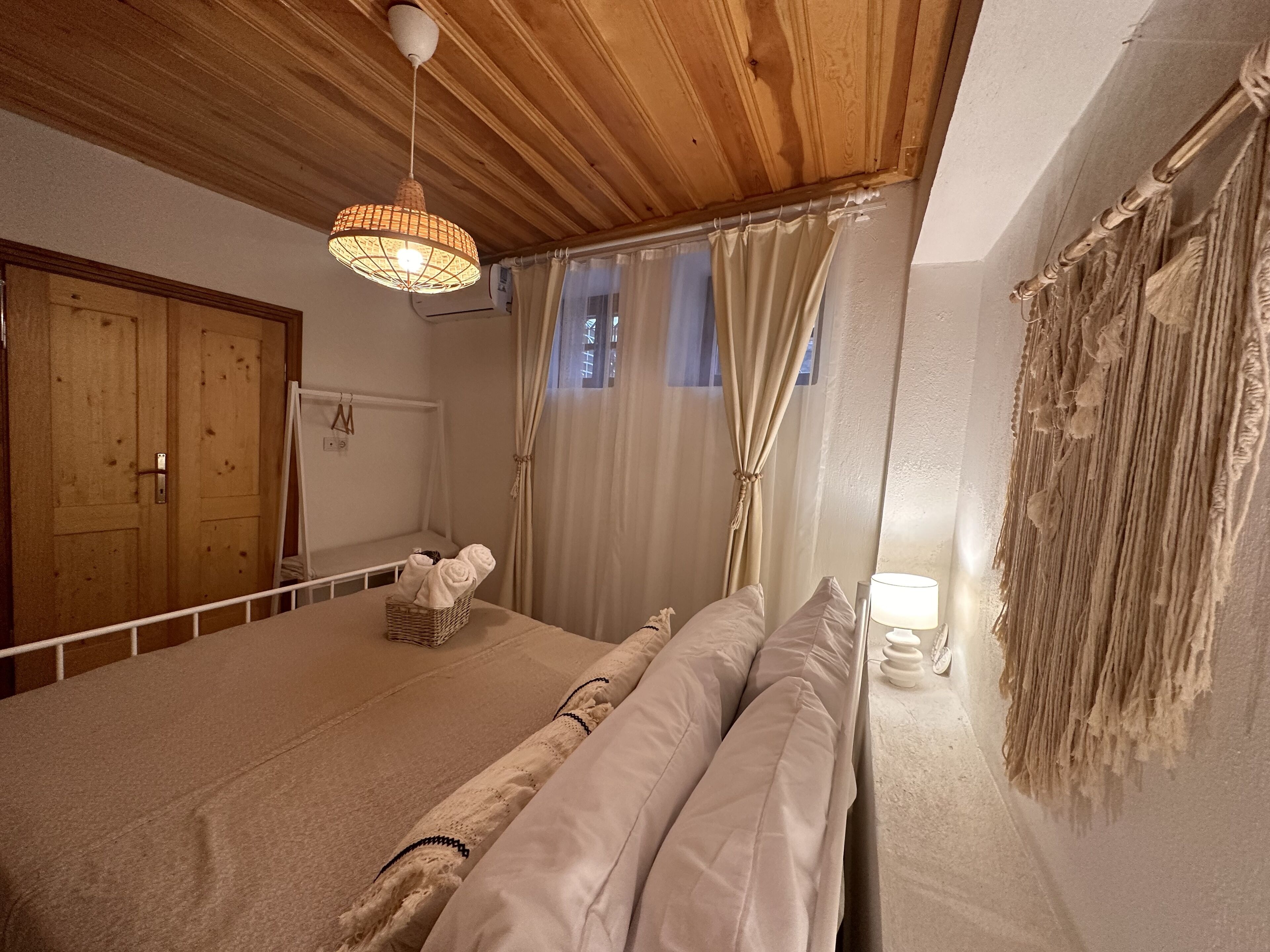 Une pièce avec un plafond en bois, un lit recouvert de draps blancs, un luminaire suspendu et une fenêtre avec des rideaux.