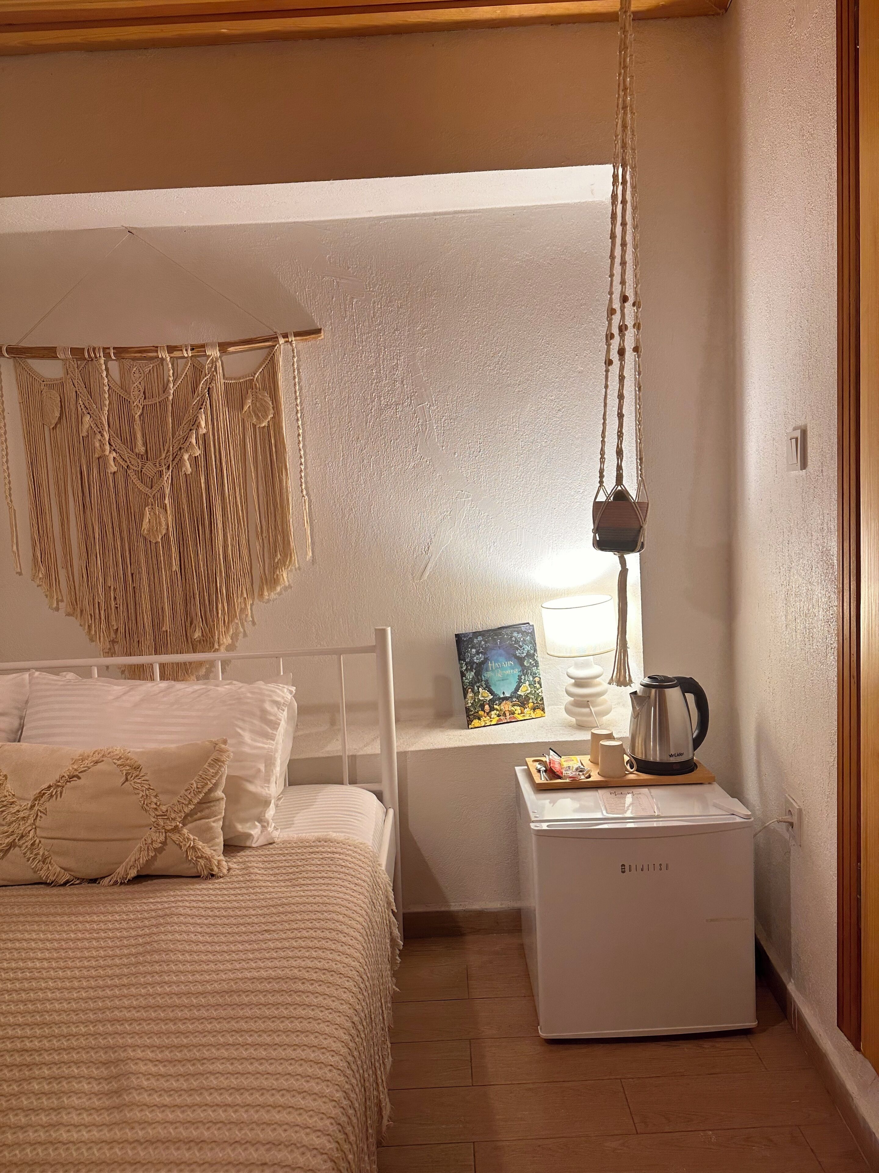 Un dormitorio con cama, un colgador de macramé en la pared, una mesita de noche con hervidor y un libro en la pared.