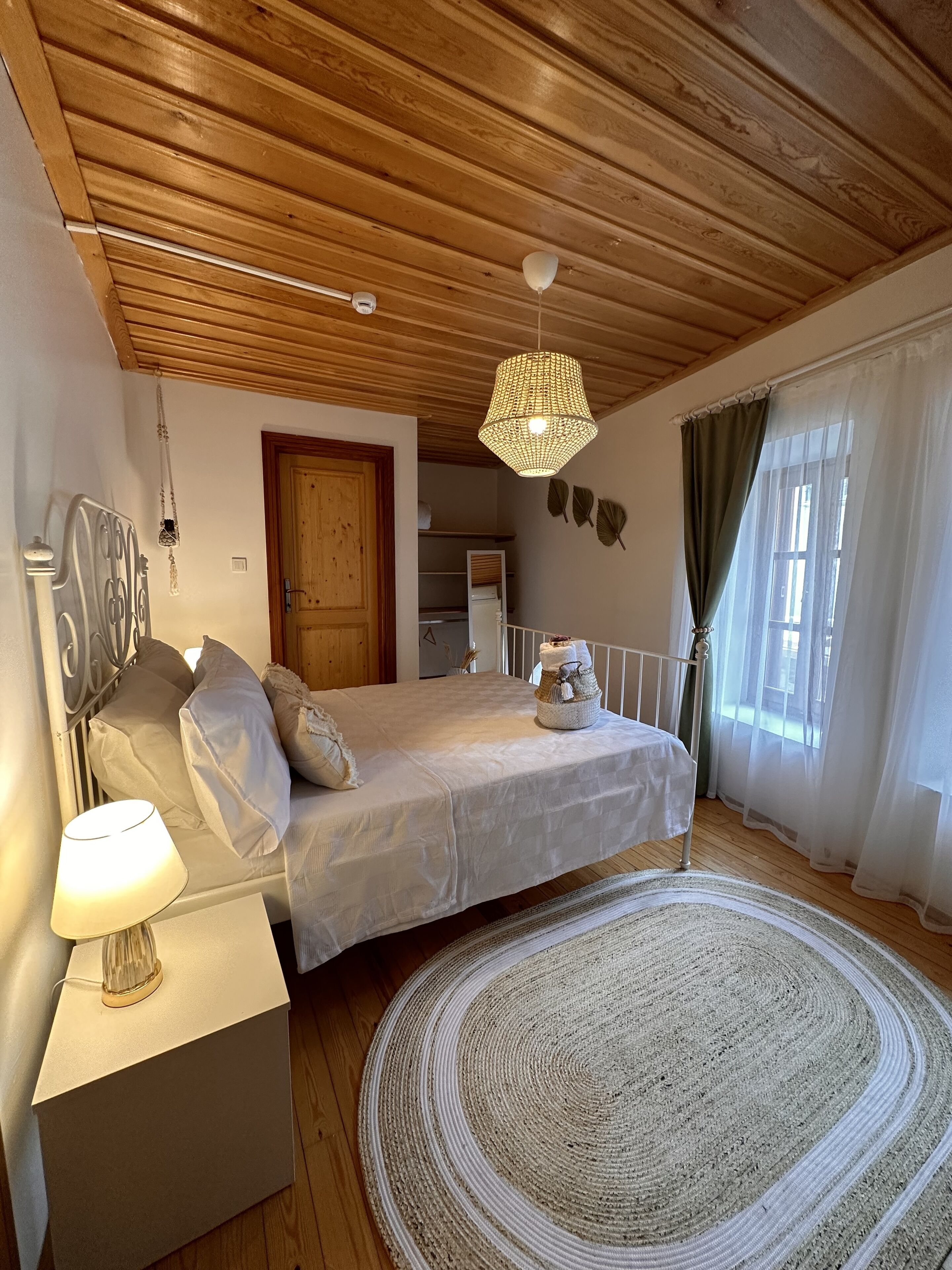 A bedroom with a wooden ceiling, a bed with a white bedspread, a bedside lamp, a woven rug, and a window with sheer curtains.