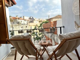 Un balcón con dos sillas de madera, una mesa pequeña con una tetera y tazas, y vistas de edificios al fondo.