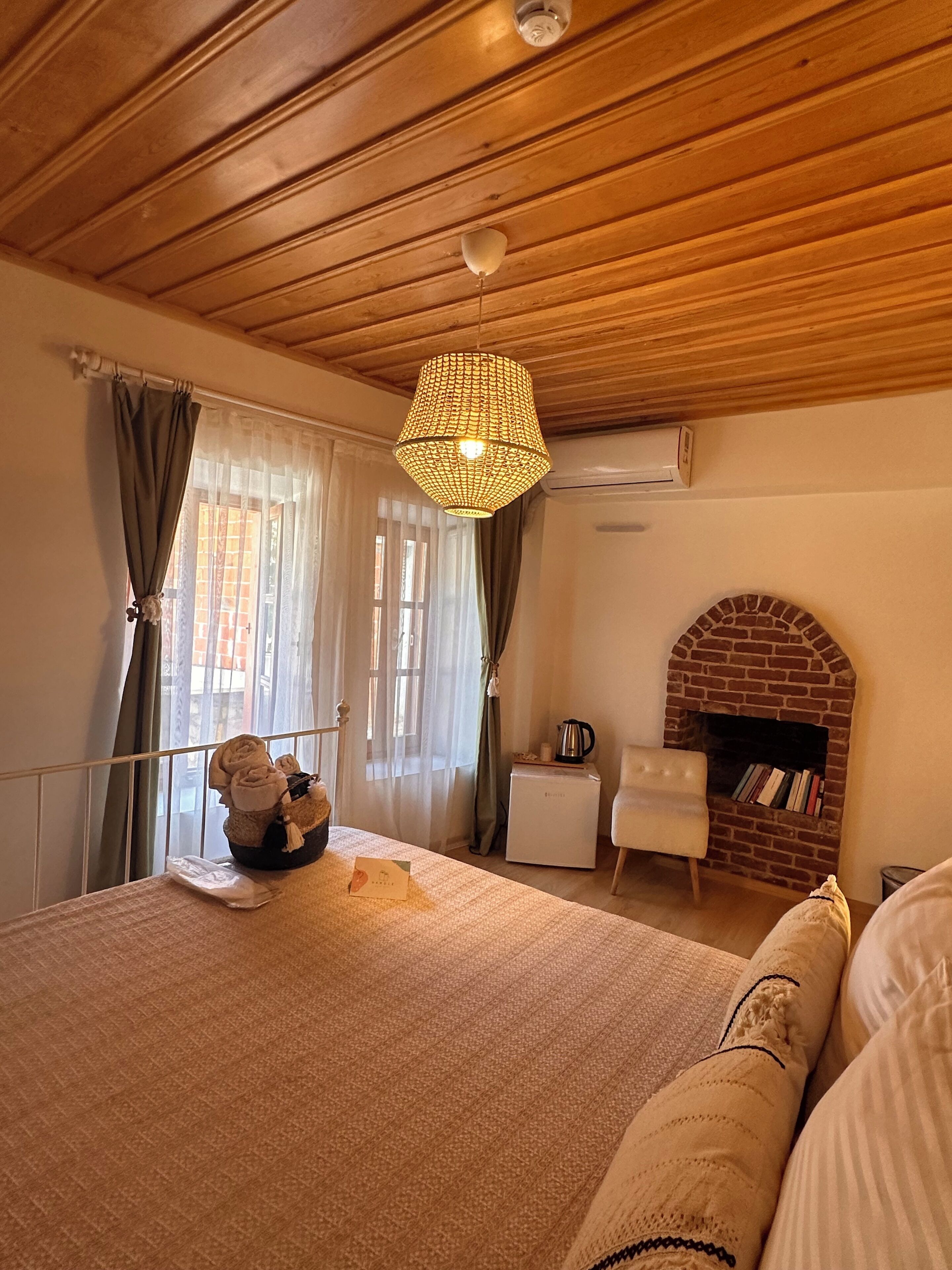 Un dormitorio con techo de madera, lámpara colgante, chimenea de ladrillo, una silla blanca y una mesita de noche con hervidor.