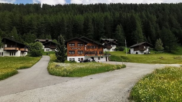 A mountain lodge with a gravel driveway, surrounded by lush greenery and a forested backdrop. - Pension Lärch (Obergoms)