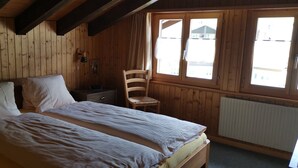 A wooden-paneled room with a bed, a chair, a nightstand, and a window with curtains. - Pension Lärch (Obergoms)