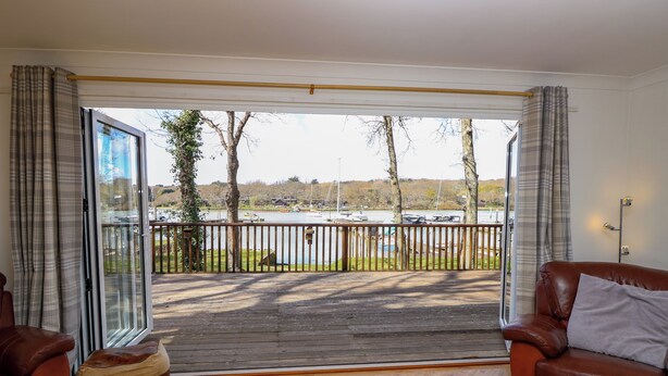 A spacious living room with a wooden floor, two leather sofas, a wooden coffee table, and a large sliding glass door leading to a balcony with a view of a lake and boats. - Byewater (Ryde)