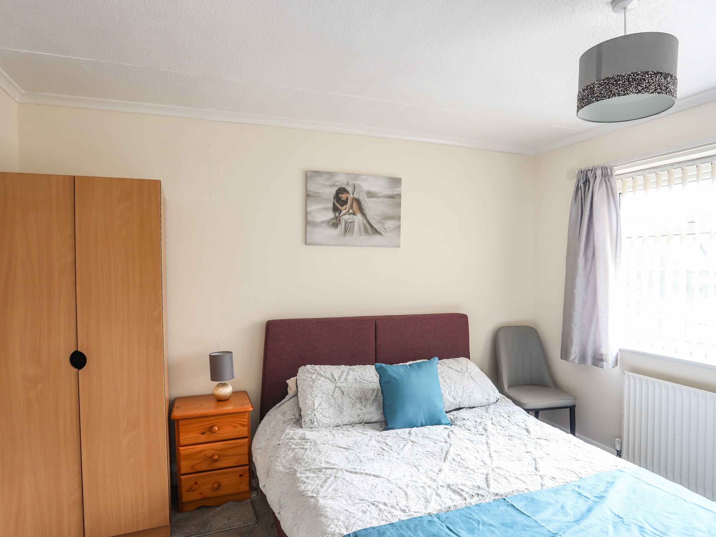 A bedroom with a bed, a wardrobe, a chair, a desk, a lamp, a painting, and a window with curtains.