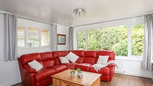 A living room with a red leather sectional sofa, a wooden coffee table, and a view of greenery through large windows. - Haddef Penmaenmawr (Penmaenmawr)