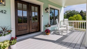 A porch with a double wooden door, potted plants, and white Adirondack chairs.