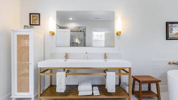 Ein modernes Badezimmer mit einem weißen Waschtisch, zwei Waschbecken, einem großen Spiegel und einem hölzernen Hocker.