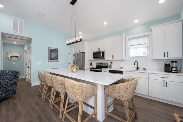 A modern kitchen with white cabinets, a central island with bar stools, stainless steel appliances, and a hanging light fixture. - Walk to the beach, Dogs ok! Huge SW community Pool -Close to all! (Corolla)
