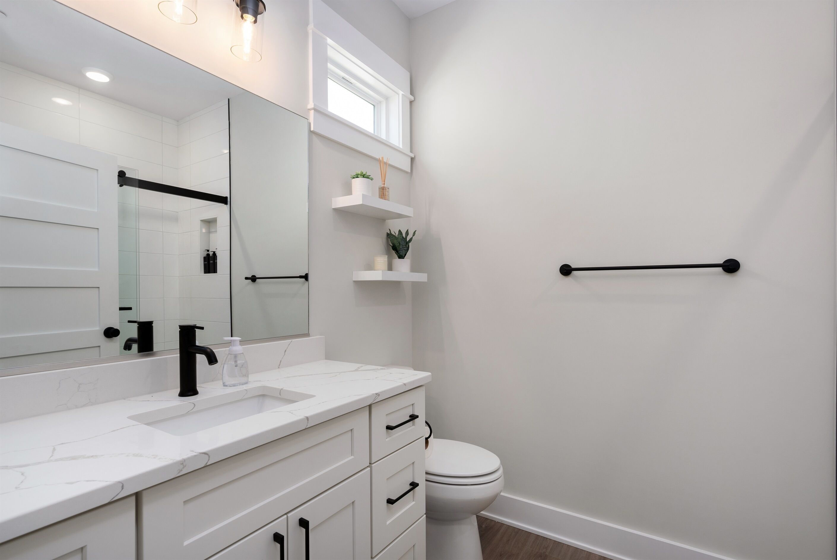 A modern bathroom with a white vanity, a large mirror, a toilet, and a small window.