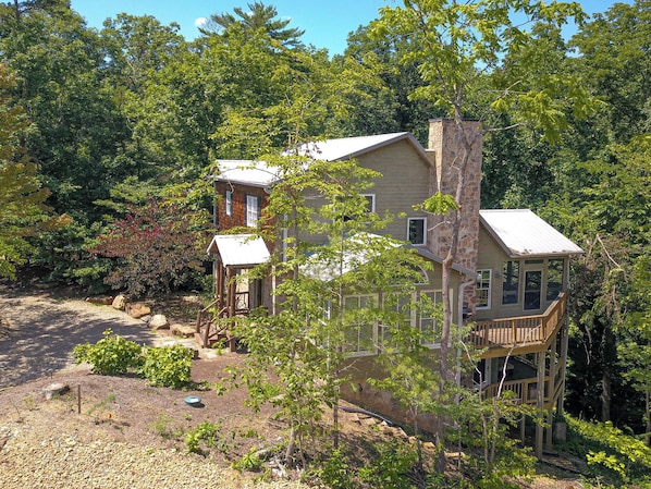 A two-story house with a stone chimney, surrounded by dense trees and a gravel driveway.