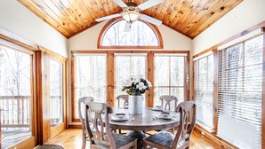 Un comedor con mesa y sillas de madera, un jarrón de flores y amplias ventanas con persianas.