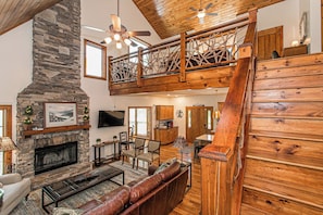 A spacious living area with a stone fireplace, a wooden staircase, and a loft with a balcony.