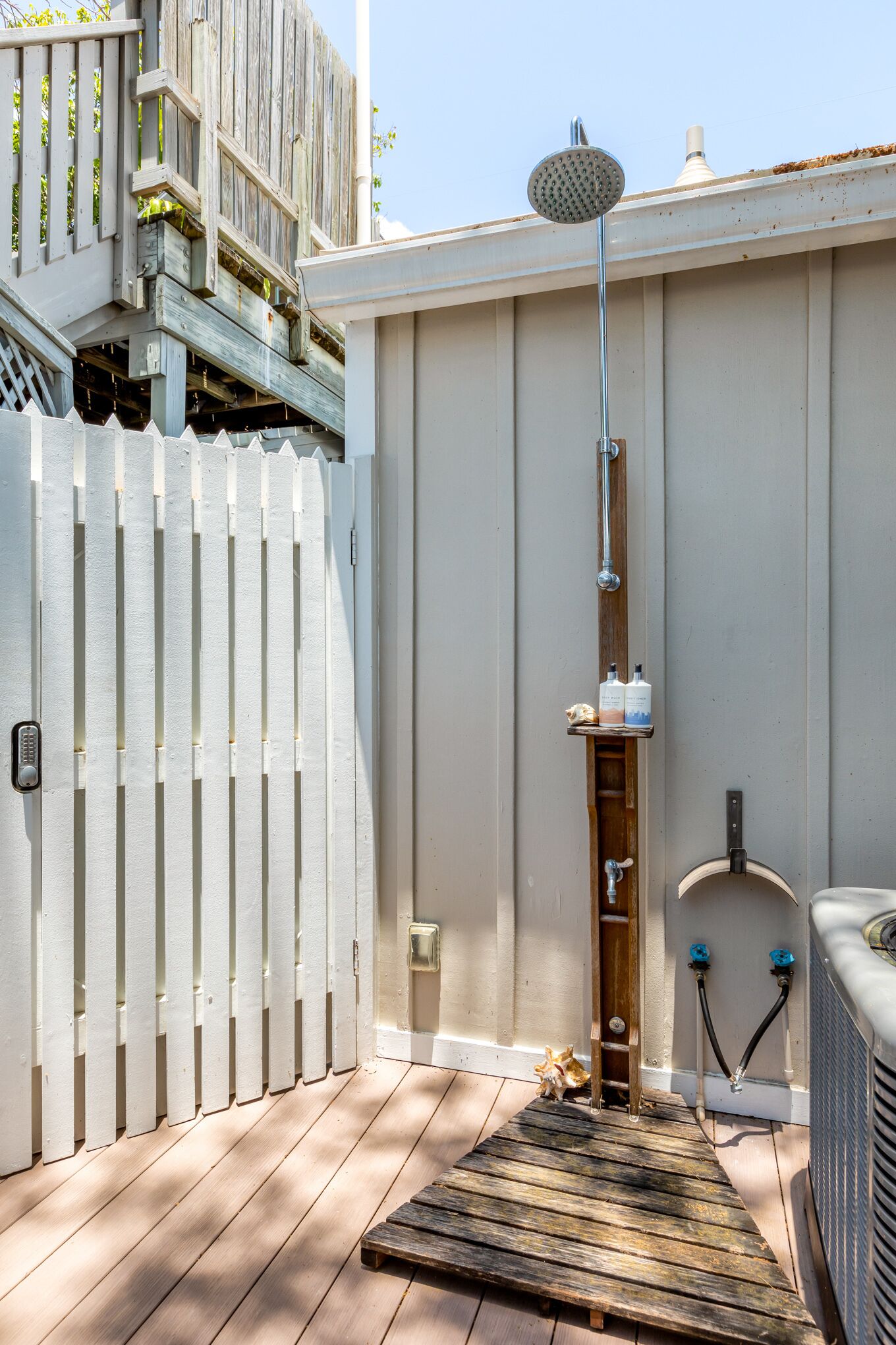 A wooden deck with a metal showerhead, a white picket fence, and a small shelf with bottles.