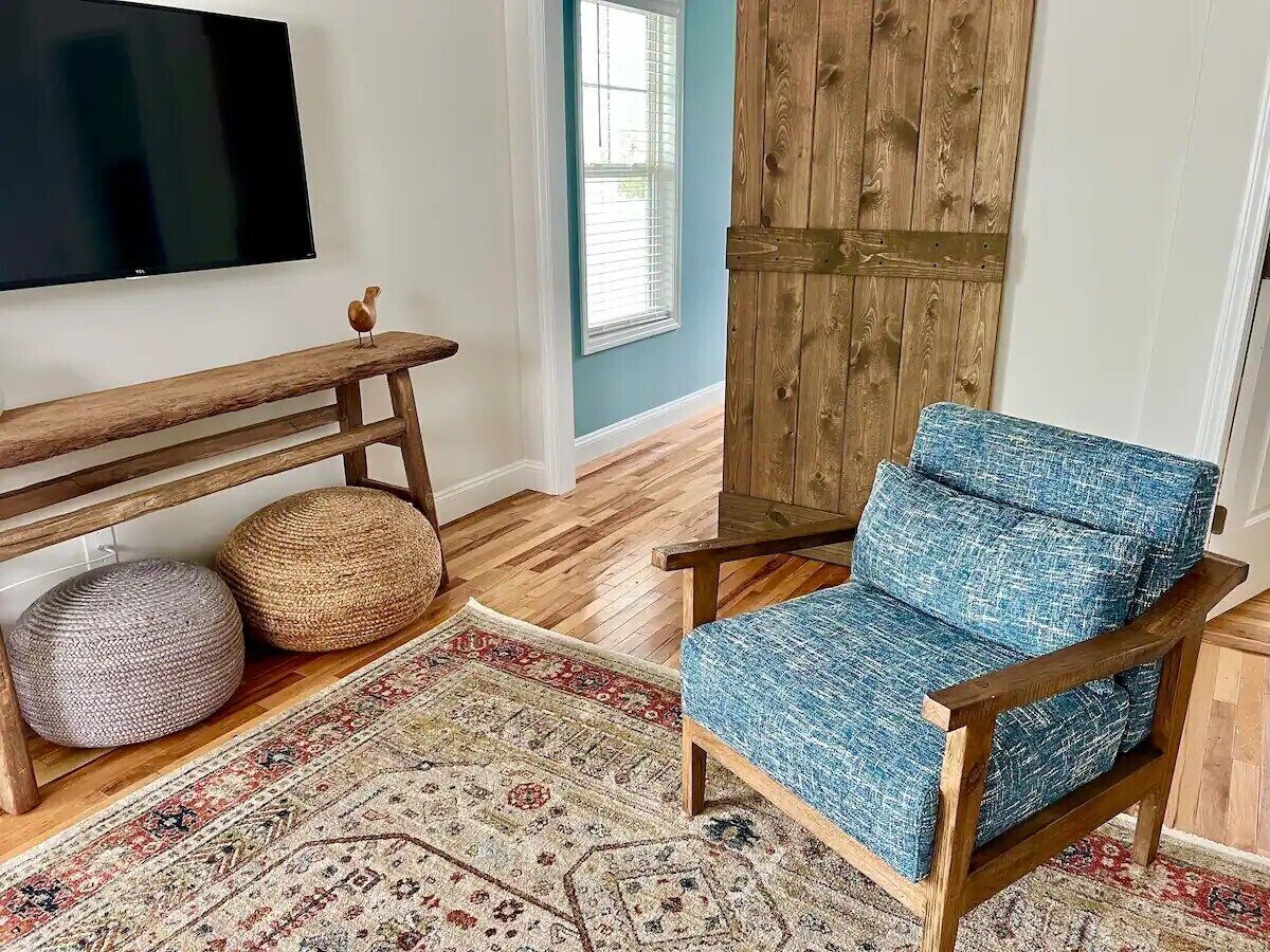 Een woonkamer met een blauwe fauteuil, een houten bankje, twee ronde poufs, een kamerkleed met een patroon en een aan de muur bevestigde televisie.
