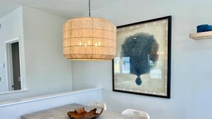 Een eetgedeelte met een licht houten tafel, witte stoelen, een geweven hanglamp en een abstract schilderij in een lijst aan de muur.