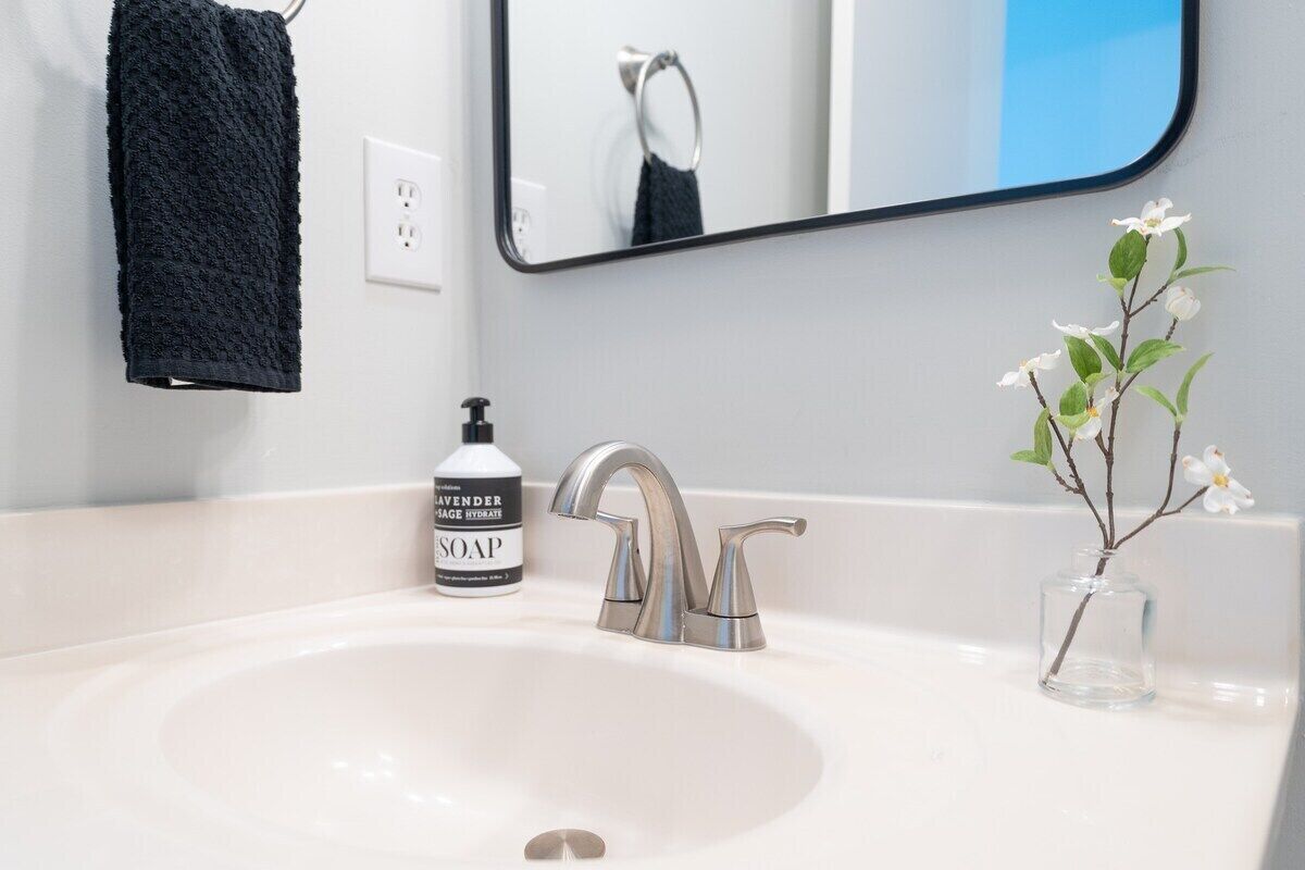 Ein Badezimmerwaschbecken mit modernem Wasserhahn, ein schwarzes Handtuch an der Wand, eine Seifenflasche und ein kleiner Blumenstrauß mit weißen Blumen.
