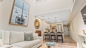 A modern living room with a sofa, a coffee table, and a painting of boats. There is a kitchen in the background with a bar and stools. - Chic Downtown Loft - Parking - Central New (Wilmington)