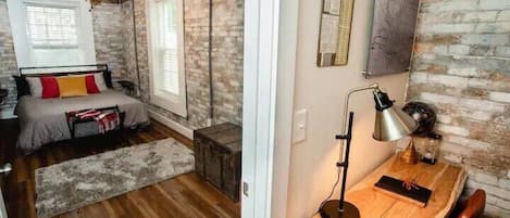 A bedroom with a bed, a desk, a chair, a lamp, and a brick wall.