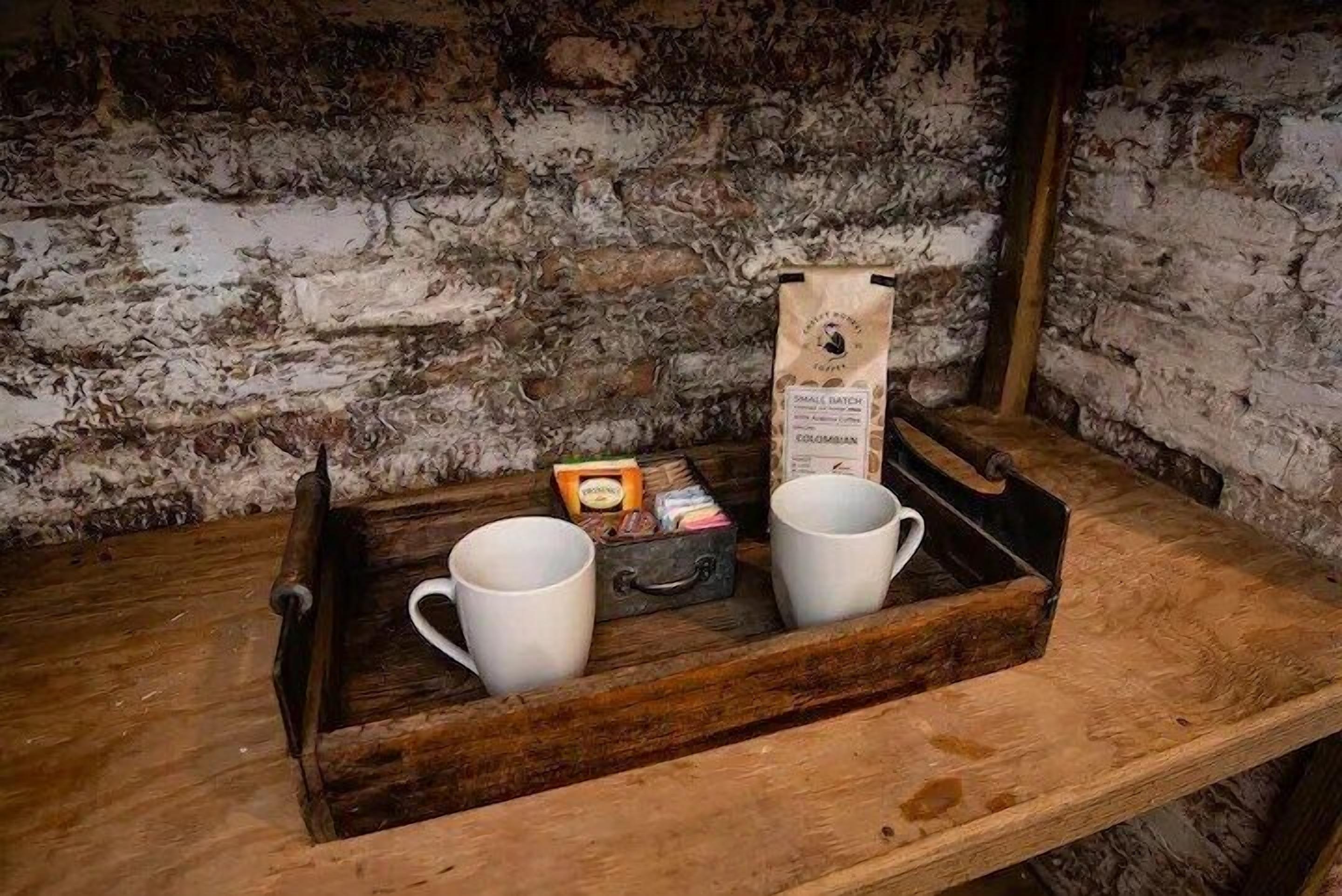 A rustic wooden tray with two white mugs and a small box of tea, placed on a wooden shelf against a textured brick wall.