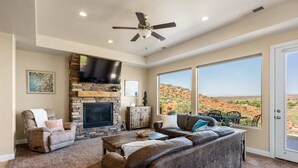 A spacious living room with a stone fireplace, comfortable seating, a flat-screen TV, and a large window offering a view of a desert landscape. - Gateway to Fun: Private Hot Tub, Community Pools, Bbq (Washington)