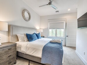 A bedroom with a bed, a nightstand, a dresser, a ceiling fan, a window with a view, and a door.