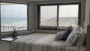 Ein Schlafzimmer mit Bett, zwei Fenstern mit Jalousien, einem kleinen Nachttisch mit Pflanze und Blick auf die Stadt.