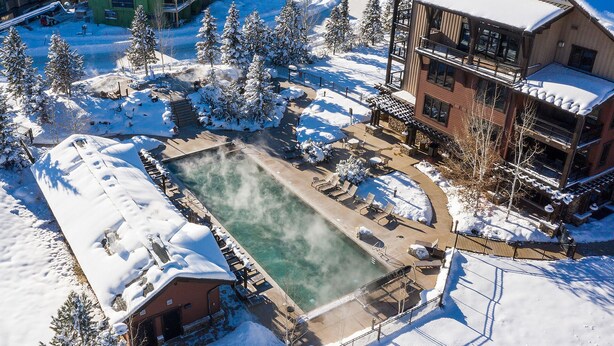 Area piscina esterna innevata con sdraio, circondata da edifici e alberi con neve.