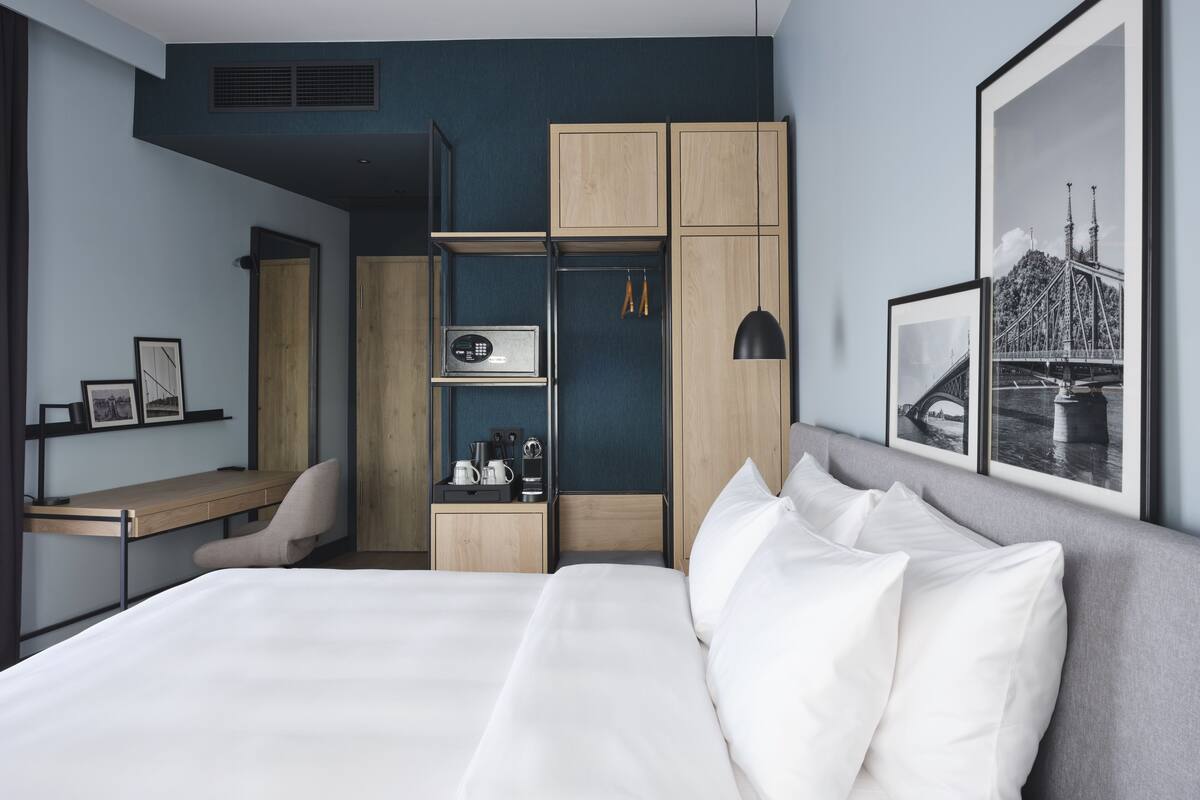 A modern hotel room with a bed, a desk, a wardrobe, and a framed picture of a bridge.