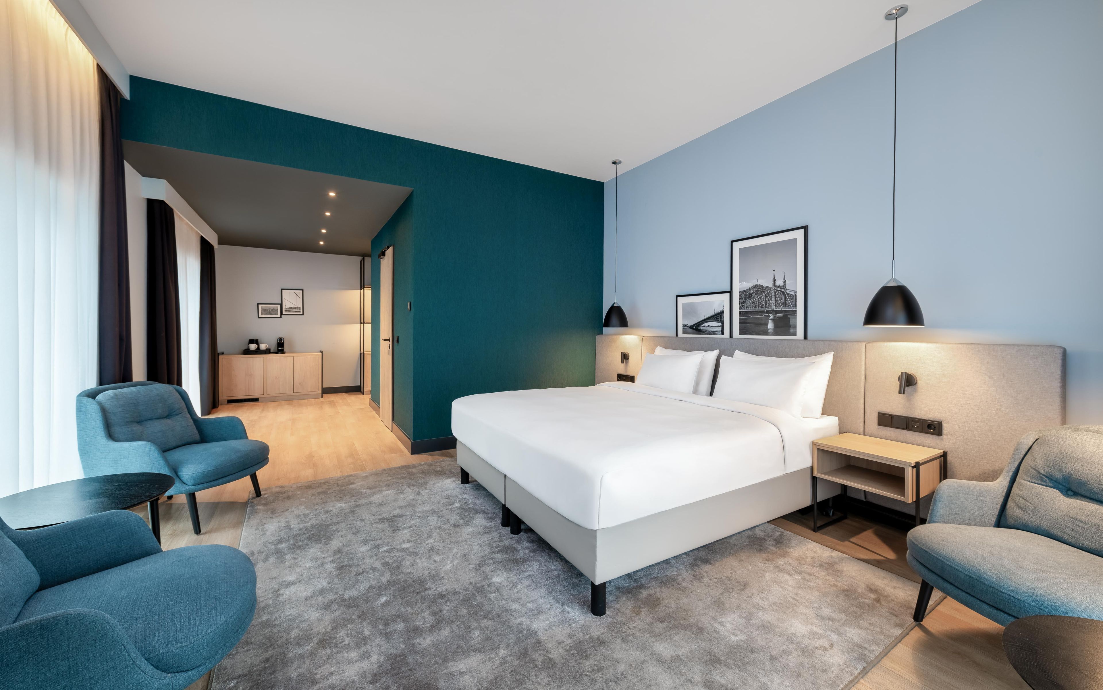 A modern hotel room with a large bed, a desk, two chairs, a wardrobe, and a bathroom visible through an open door.