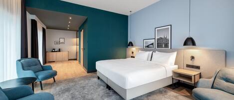 A modern hotel room with a large bed, a desk, two chairs, a wardrobe, and a bathroom visible through an open door.