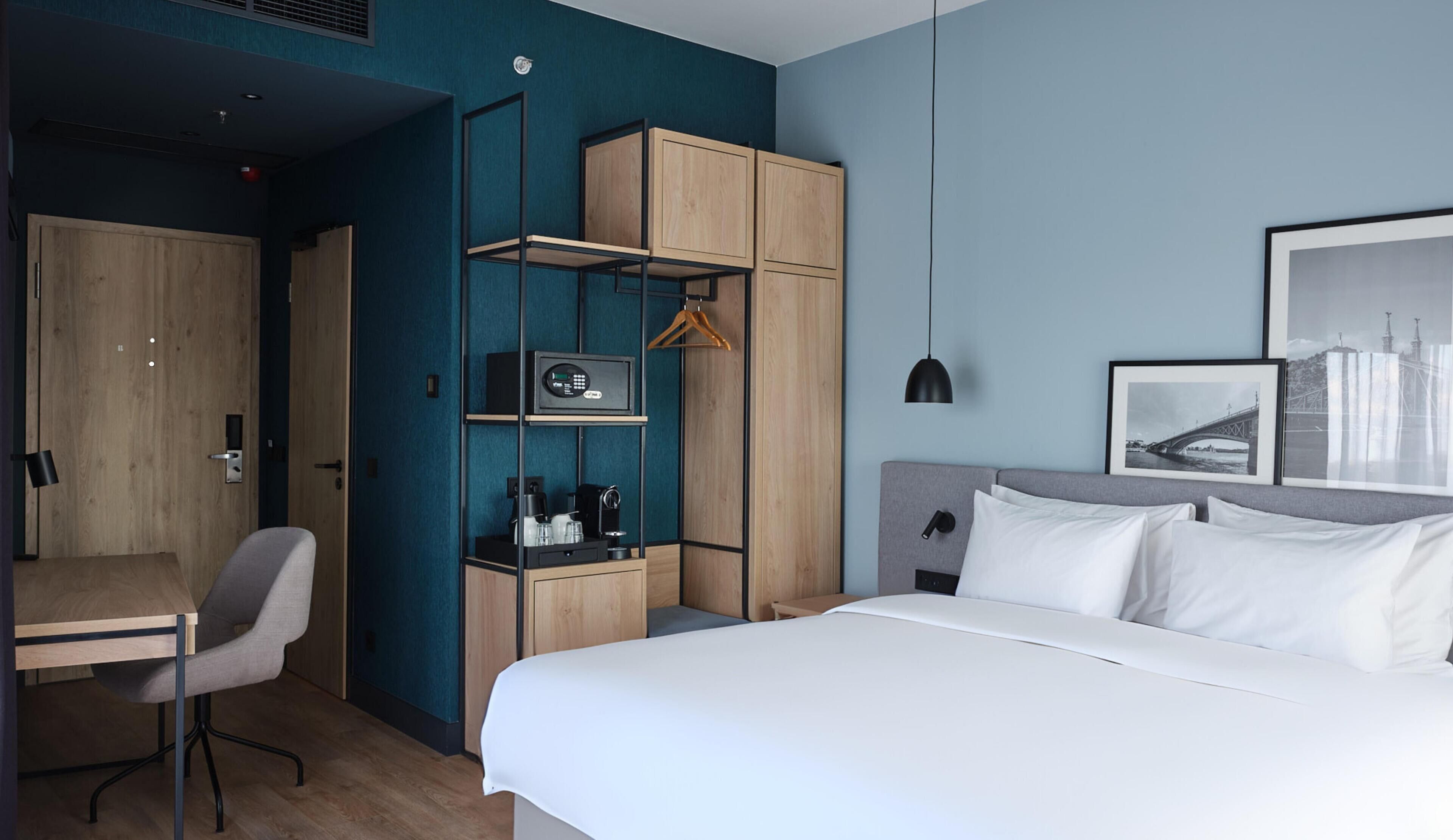 A modern hotel room with a bed, a desk, a chair, a wardrobe, and a hanging coat rack.