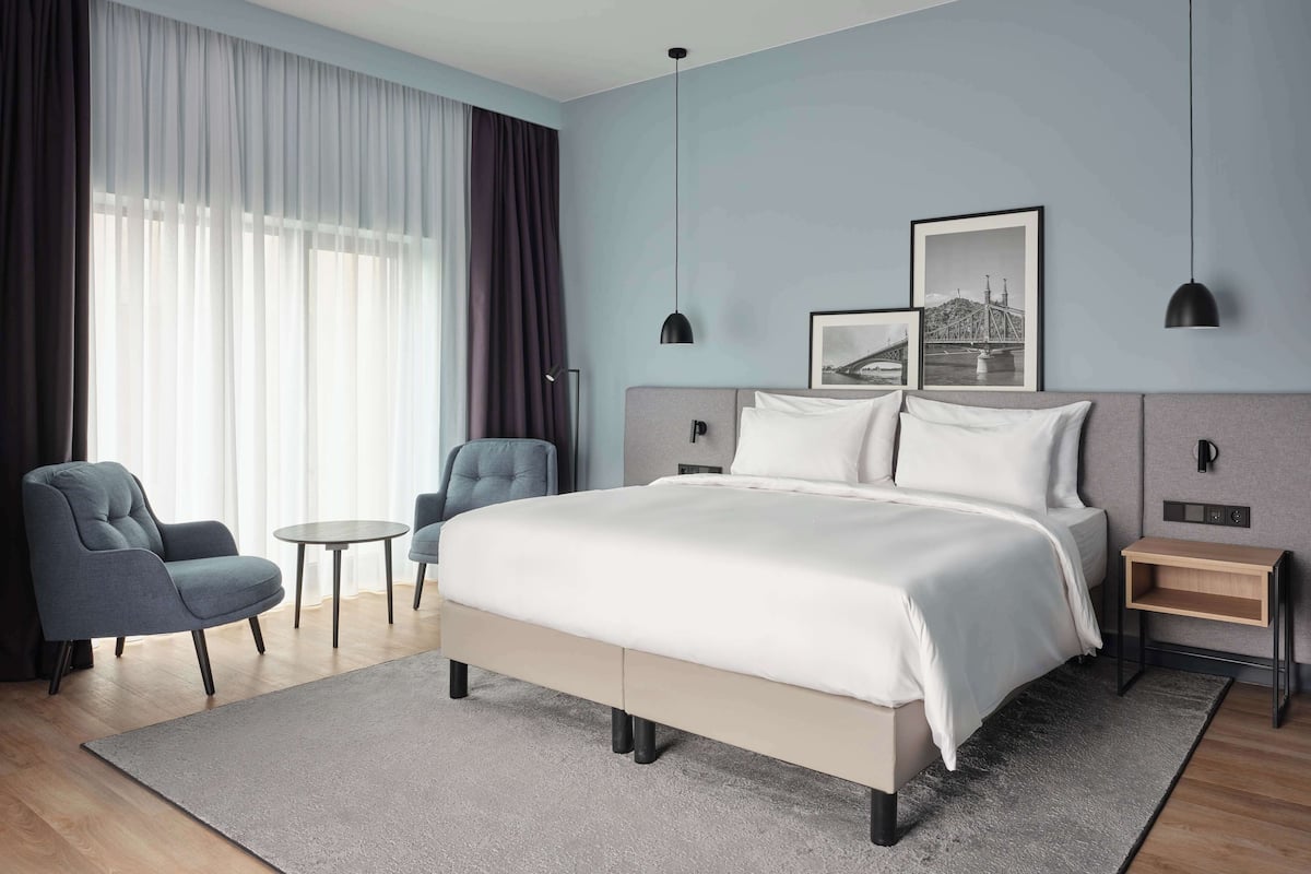A modern hotel room with a large bed, two armchairs, a small table, and a nightstand.