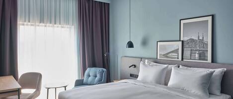 A modern hotel room with a large bed, a desk, a chair, and two framed pictures on the wall.