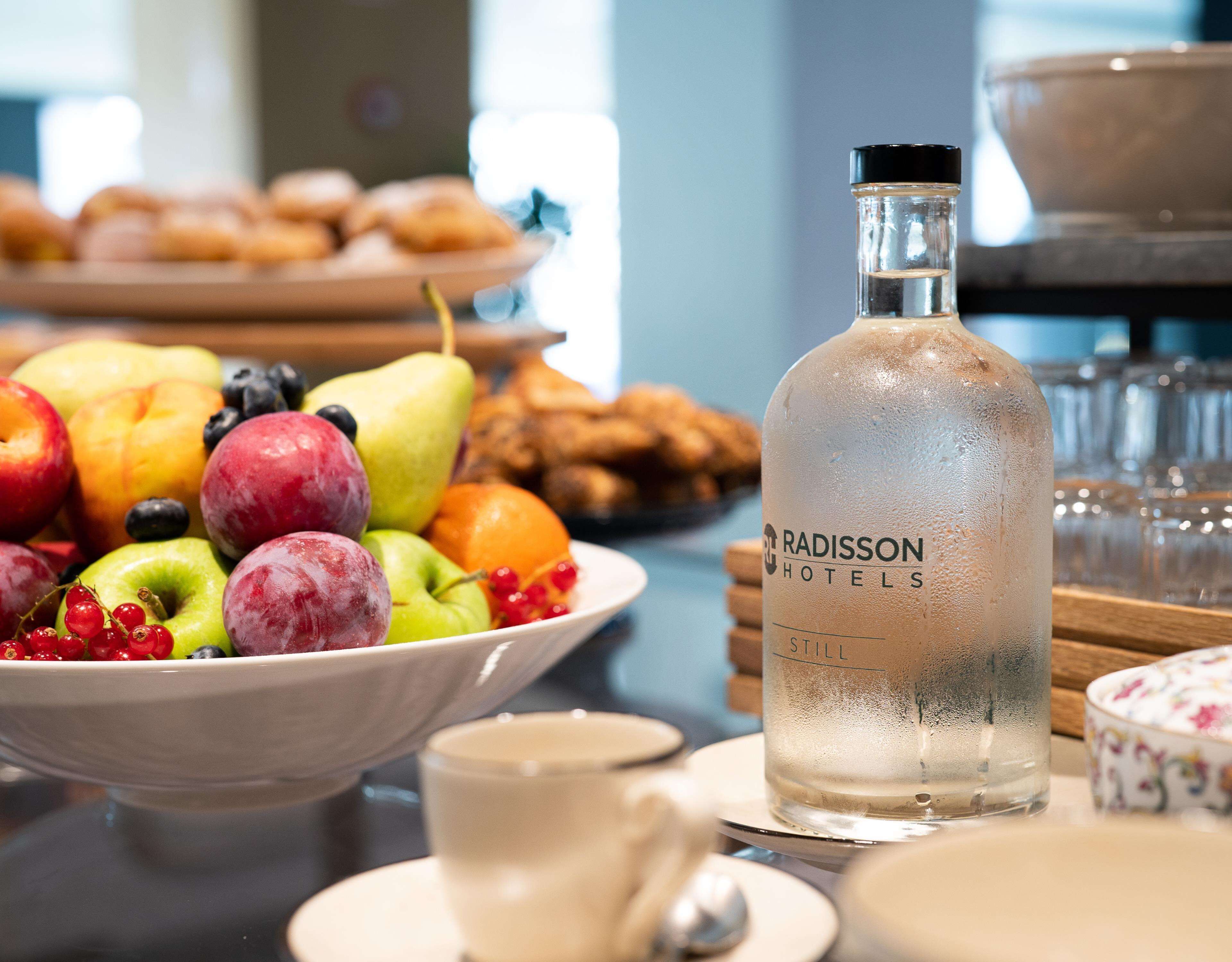 A glass bottle labeled "RADISSON HOTELS" with a still water bottle beside a bowl of assorted fruits and pastries.
