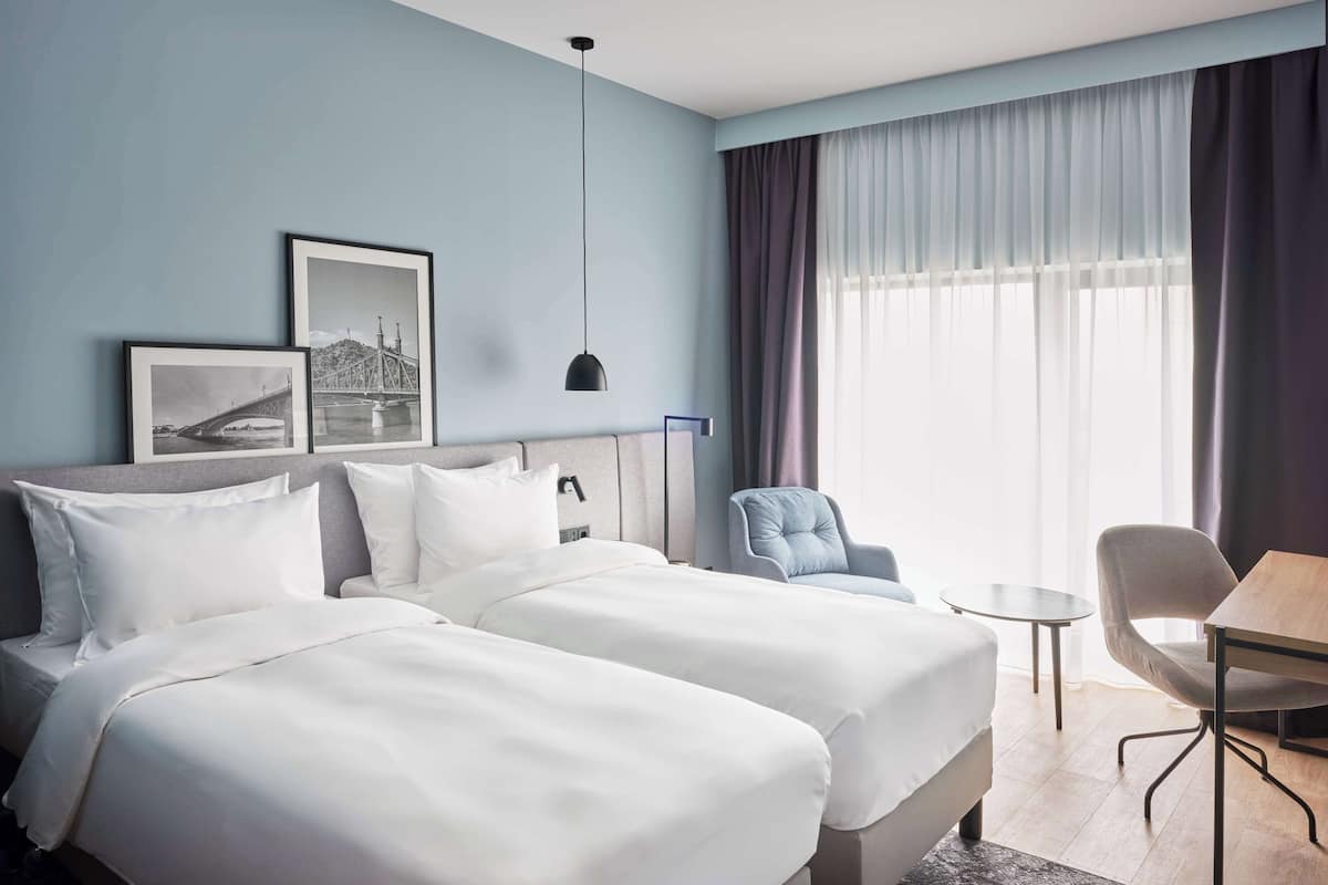 A hotel room with two beds, a desk, a chair, and a large window with curtains.