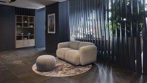 A modern interior with a sectional sofa, a round ottoman, a wooden shelving unit, and a large window with vertical blinds. - Radisson Hotel Budapest Budapart (Budapest)