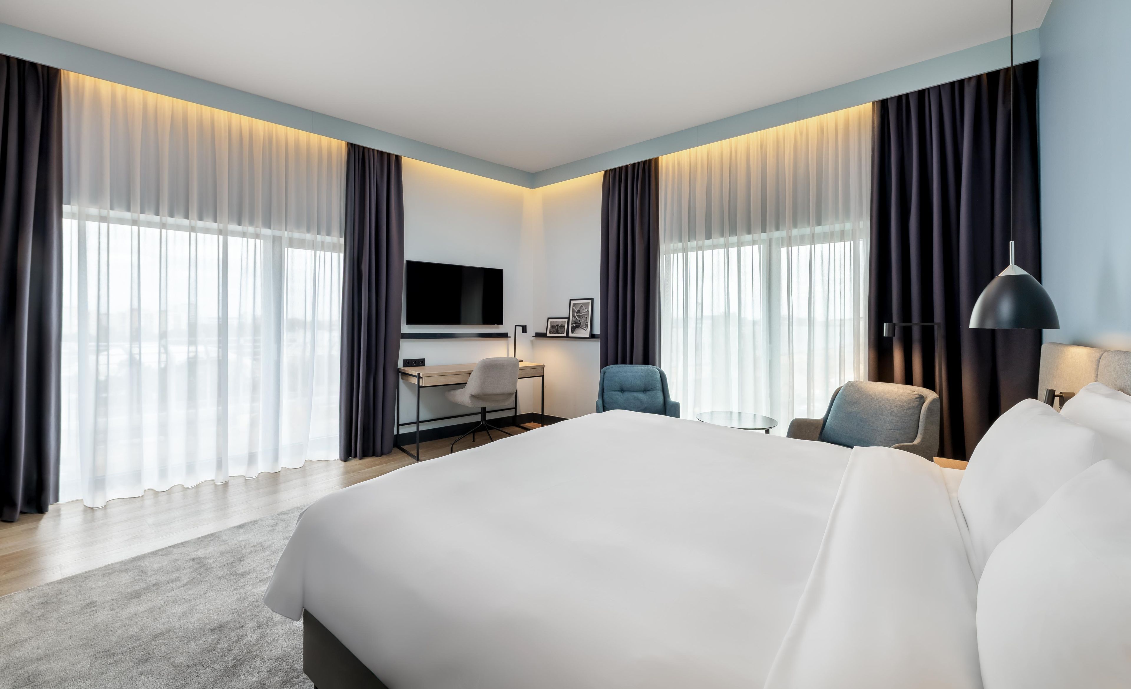 A modern hotel room with a large bed, a desk, a chair, and a TV.