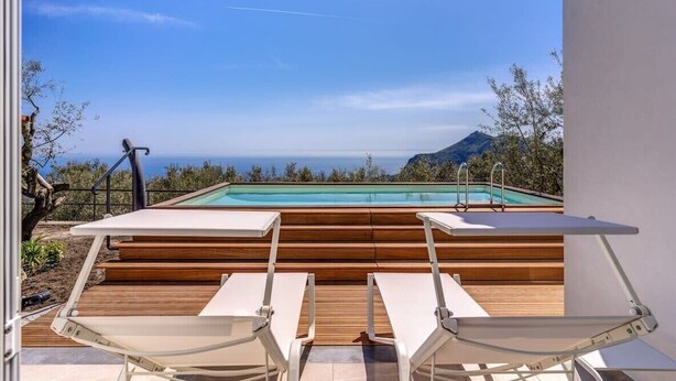 Ein Poolbereich mit Holzstufen zum Pool, umgeben von weißen Liegestühlen und freiem Blick auf das Meer und die Berge.