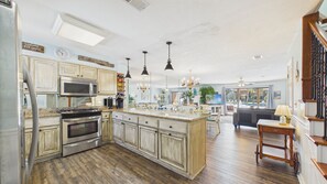 Una cocina con armarios de madera, una isla central, electrodomésticos de acero inoxidable, iluminación colgante y vistas al área de estar.