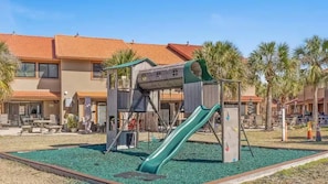 A playground with a slide and climbing structures, surrounded by palm trees and a building with a tiled roof. - Paradise Pointe 137–5 Min to Beach–Sleeps 6 (Panama City Beach)